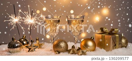 Elegant Christmas celebration scene with sparkling wine glasses, golden baubles, sparklers, and a gift box in festive snow setting 130209577