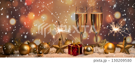 Christmas celebration with champagne glasses, sparklers, golden baubles, and red gift on snow with warm festive background 130209580