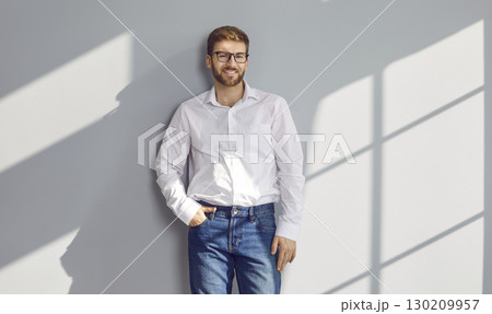 Portrait of man in white shirt, blue jeans and glasses standing against light grey wall 130209957