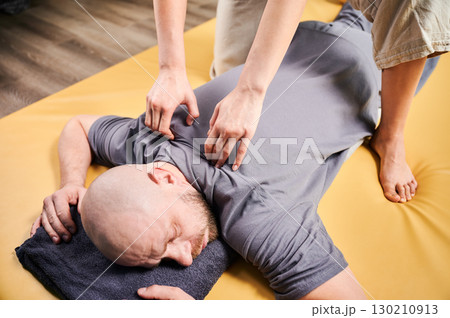 Masseuse making thai yoga massage. Therapist doing traditional massage treatment for male patient indoors. Bald client lying on mat, meditating, enjoying hand massage in calm atmosphere. 130210913