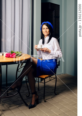 A woman sits comfortably at a small round table, holding a cup of coffee A woman sits comfortably at a small round table, holding a cup of coffee 130212000