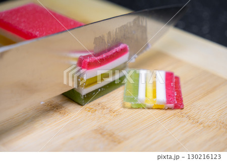 A sharp kitchen knife precisely slicing vibrant multi-layered jelly candy block on a bamboo cutting board, preparing colorful festive sweet dessert 130216123