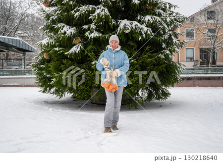 Woman in blue winter coat walking in snowy park with mesh bag of tangerines Woman in blue winter coat walking in snowy park with mesh bag of tangerines 130218640