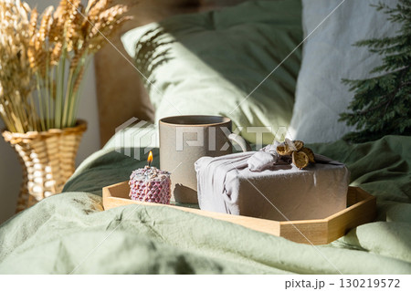 Cozy bed scene with mug, lit candle and furoshiki gift on wooden tray. Cozy bed scene with mug, lit candle and furoshiki gift on wooden tray. 130219572