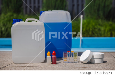 Set of pool maintenance supplies including chemical canisters, chlorine tablets, dropper bottles, and a water test kit placed near a swimming pool in sunlight. Set of pool maintenance supplies including chemical canisters, chlorine tablets, dropper bottles, and a water test kit placed near a swimming pool in sunlight. 130219974