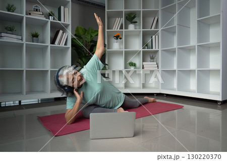 Senior woman engaging in yoga with headphones and laptop in a modern wellness space 130220370