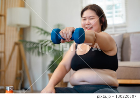 Plus-size woman lifting a dumbbell with a smile, promoting strength and healthy lifestyle at home 130220507