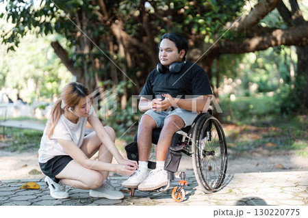 Caring friend helping a person in a wheelchair tie shoes in a serene park environment 130220775