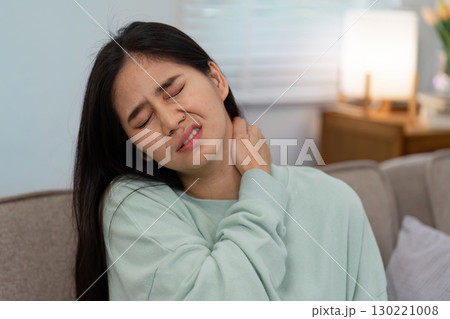 Woman in a mint green sweater grimacing in pain at home, clutching her neck to relieve discomfort and express distress Woman in a mint green sweater grimacing in pain at home, clutching her neck to relieve discomfort and express distress 130221008