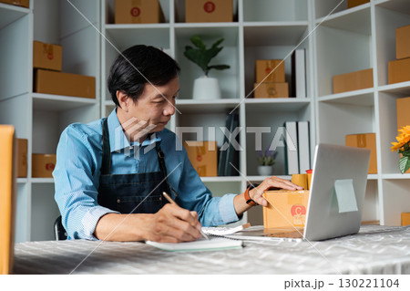 Elderly man writing notes while organizing packages in a modern workspace 130221104