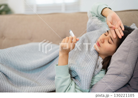 Woman in a mint green sweater lying on a sofa at home, checking her temperature with a thermometer, exhibiting signs of sickness 130221105