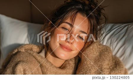 Portrait of a young woman wrapped in a soft wool blanket, reclining on a bed with plush pillows in gentle, warm indoor lighting 130224805