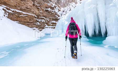 Female hiker with backpack and trekking poles exploring a frozen canyon with stunning ice formations and turquoise water, enjoying the tranquility of winter nature 130225063