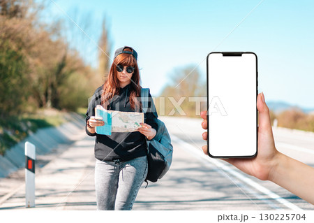 Hitchhiking and local travel. A young woman in cap and sunglasses stand along the road with a paper map. Female hands holding a smartphone with mock up. Copy space. Concept of online guide 130225704