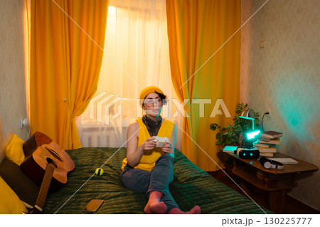 Teen girl enjoying peaceful solo gaming moment in her room. Sitting comfortably on bed, surrounded by warm light. Concept of digital comfort and fun. Wide shot 130225777