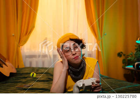 Teen girl in relaxed posture gaming at home in evening light. Cozy environment with guitar and warm pillows. Concept of youth freedom and tech. Mid shot 130225780