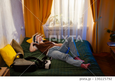 Woman sits on bed in softly lit room holding VR glasses away from face, showing discomfort. Realistic reaction to digital interaction and new tech exploration. Wide shot 130225792