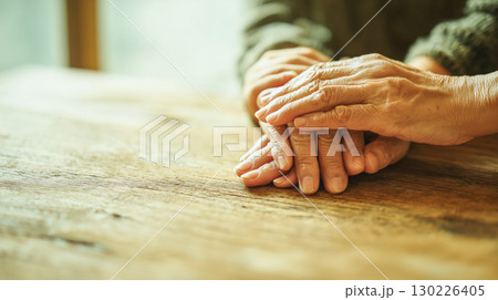 温かな光の室内、木製テーブルの上で重なる高齢者の手—優しさと安らぎを感じる瞬間 130226405