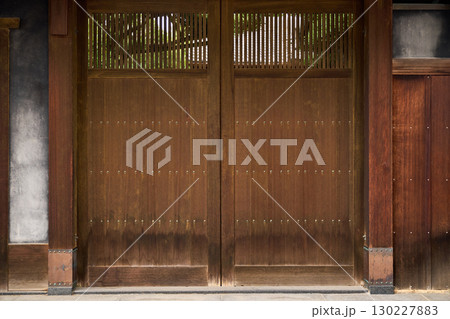 伝統的な日本建築の背景用素材。落ち着いた風合いの木製の門扉と格子から透けて見える家屋と木。 伝統的な日本建築の背景用素材。落ち着いた風合いの木製の門扉と格子から透けて見える家屋と木。 130227883