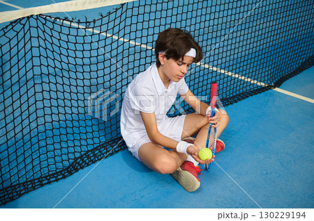 Young Tennis Player Sitting Beneath the Net Holding a Tennis Ball Young Tennis Player Sitting Beneath the Net Holding a Tennis Ball 130229194