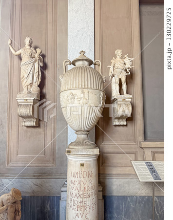VATICAN CITY, ROME, ITALY - AUGUST 02, 2025 Classical marble vase with fluted body and handles sits on an inscribed pedestal. Two smaller statues are on ornate brackets flanking the central urn VATICAN CITY, ROME, ITALY - AUGUST 02, 2025 Classical marble vase with fluted body and handles sits on an inscribed pedestal. Two smaller statues are on ornate brackets flanking the central urn 130229725