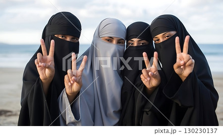 Four women wearing hijab making peace signs on a beach outdoors smiling 130230137