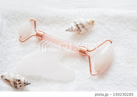 Rose quartz facial roller and gua sha stone arranged on a soft white towel with decorative seashells for a skincare and beauty spa concept Rose quartz facial roller and gua sha stone arranged on a soft white towel with decorative seashells for a skincare and beauty spa concept 130230285
