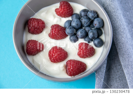 Natural fresh greek yogurt with red raspberry, blue berries in the grey plate and napkin for healthy breakfast. Blue paper background.  130230478