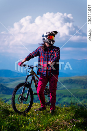Unrecognizable guy riding a mountain bike on top of a mountain. Young man with a bicycle standing in a green field against the backdrop of mountains and cumulus clouds, vertical photo 130231047