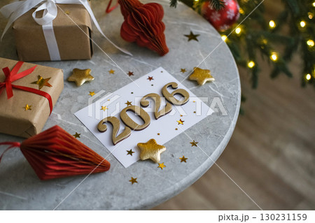 Greeting card with gold numbers 2026 and stars surrounded by gift boxes and holiday decorations on round marble table near Christmas tree 130231159