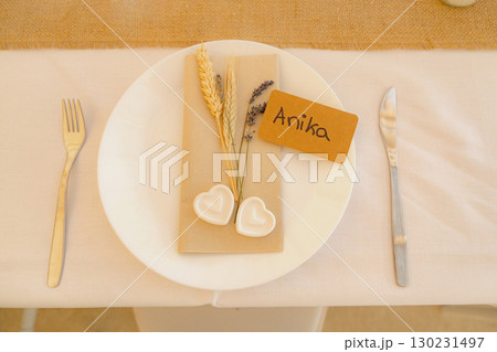 Elegant table setting featuring a name card, decorative wheat, and heart-shaped adornments on a plate. Knife and fork are neatly placed, evoking a sense of sophistication for a special occasion. 130231497