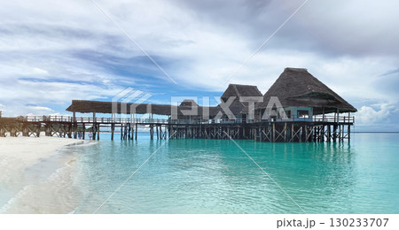 A picturesque wooden pier and restaurant with thatched roofs extend over the turquoise waters of the Indian Ocean, seen from a pristine white-sand beach 130233707