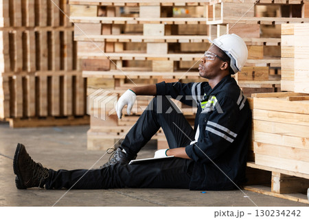 African American male worker in safety gear takes break while sitting on warehouse floor, surrounded by wooden pallets, representing industrial fatigue, rest and labor conditions. African American male worker in safety gear takes break while sitting on warehouse floor, surrounded by wooden pallets, representing industrial fatigue, rest and labor conditions. 130234242