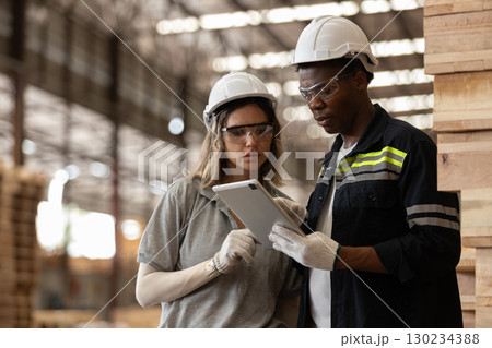 Two Multi-ethnic industrial workers wearing safety helmets and gloves use digital tablet for data analysis in warehouse, showcasing teamwork, technology wooden manufacturing processes. 130234388