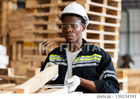 African American Male worker wearing safety gear inspects wooden planks in lumber warehouse, focusing on quality control, industrial safety, and manufacturing operations. African American Male worker wearing safety gear inspects wooden planks in lumber warehouse, focusing on quality control, industrial safety, and manufacturing operations. 130234391