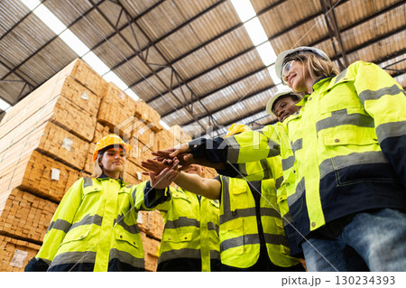 Group of wood warehouse workers in high visibility safety gear stacking hands in teamwork gesture inside lumber storage facility, symbolizing unity, motivation, and collaboration in industrial work. Group of wood warehouse workers in high visibility safety gear stacking hands in teamwork gesture inside lumber storage facility, symbolizing unity, motivation, and collaboration in industrial work. 130234393