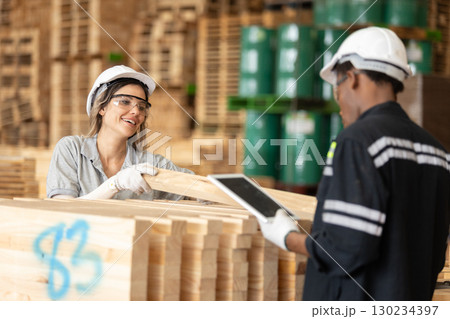 Female and male workers in safety gear collaborate in a lumber warehouse, handling wood planks and using digital tablet for inventory or quality control processes. Female and male workers in safety gear collaborate in a lumber warehouse, handling wood planks and using digital tablet for inventory or quality control processes. 130234397