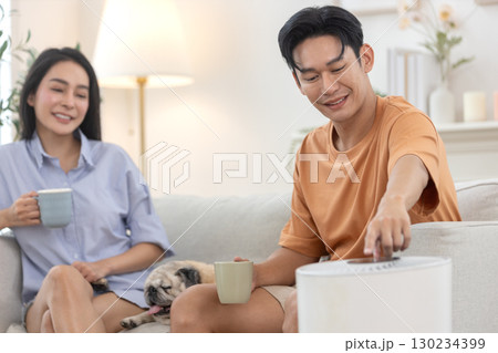 young asian couple drinking morning coffee sitting on sofa relaxing time living room with small pug dog rests beside, man pressing power button air purifier for clean air in house for good health. young asian couple drinking morning coffee sitting on sofa relaxing time living room with small pug dog rests beside, man pressing power button air purifier for clean air in house for good health. 130234399