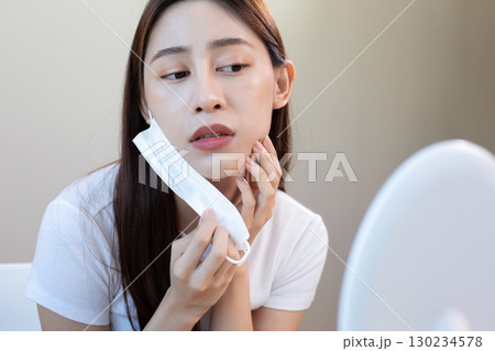 Asian woman removing face mask and checking acne in mirror with concern, representing mask problem, skincare issue, and facial irritation caused by prolonged mask use. Asian woman removing face mask and checking acne in mirror with concern, representing mask problem, skincare issue, and facial irritation caused by prolonged mask use. 130234578