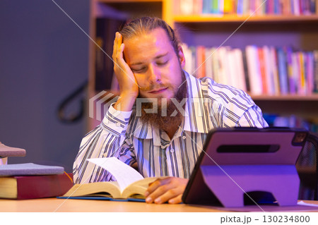 Tired man falling asleep while studying at a library with books and tablet, representing study burnout, mental fatigue, or late-night academic pressure in a learning environment. 130234800