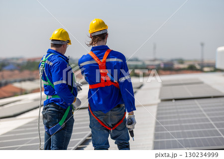Two solar panel technicians in safety gear inspecting rooftop installation site, representing renewable energy solar power systems, teamwork, clean technology industrial planning and workplace safety. 130234999