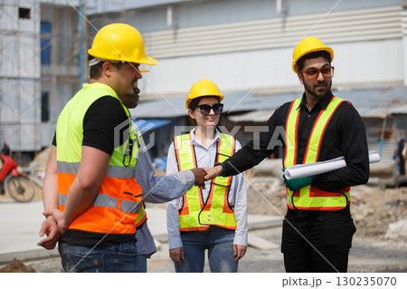 Diverse construction team wearing safety helmets and reflective vests shaking hands at building site symbolizing teamwork, agreement, partnership and successful project collaboration. Diverse construction team wearing safety helmets and reflective vests shaking hands at building site symbolizing teamwork, agreement, partnership and successful project collaboration. 130235070