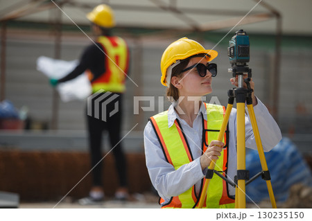 Female construction surveyor wearing safety helmet and reflective vest using leveling instrument at building site, representing measurement, engineering, precision and infrastructure development. 130235150