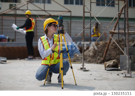 Female construction surveyor wearing safety helmet and reflective vest using leveling instrument at building site, representing measurement, engineering, precision and infrastructure development. 130235151