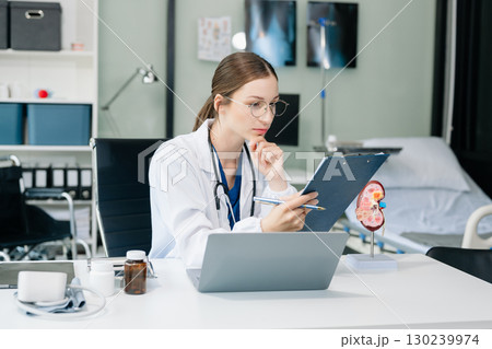 Confident young Caucasian female doctor in white medical uniform sit at desk working on computer. Smiling use laptop write in medical journal 130239974