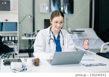 Confident young Caucasian female doctor in white medical uniform sit at desk working on computer. Smiling use laptop write in medical journal Confident young Caucasian female doctor in white medical uniform sit at desk working on computer. Smiling use laptop write in medical journal 130240541