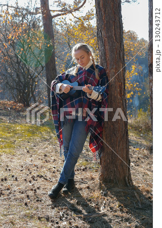 Portrait of smiling caucasian girl plays ukulele in autumnal park. Young woman plays guitar musical instrument outside in nature fall time. Audio music healing Portrait of smiling caucasian girl plays ukulele in autumnal park. Young woman plays guitar musical instrument outside in nature fall time. Audio music healing 130243712