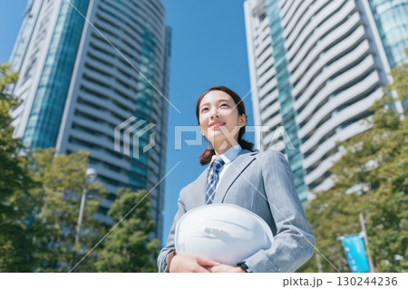 a japanese female engineer wearing a blue suit and holding a white helmet stands in front of tall office buildings 130244236