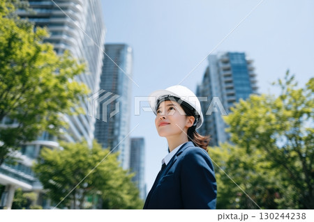 a japanese female engineer wearing a blue suit and holding a white helmet stands in front of tall office buildings 130244238