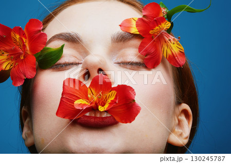 Close up portrait of a model with flowers covering her lips, vibrant colors, expressing beauty and femininity, with a blue background, perfect for concepts of wellness and nature Close up portrait of a model with flowers covering her lips, vibrant colors, expressing beauty and femininity, with a blue background, perfect for concepts of wellness and nature 130245787